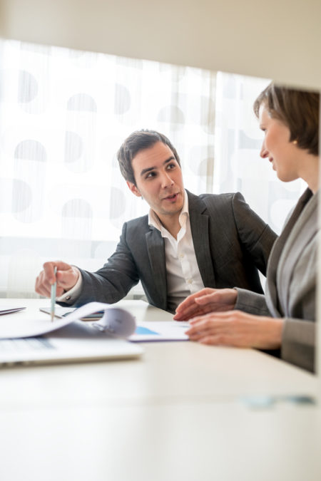 Two Young Business People Discussing at the Table Two Young Business People in Corporate Attire Discussing Some Plans at the Table with Plenty of Documents.